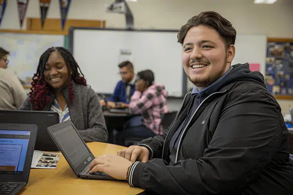 Student smiles away from his computer at the DSF Future Center. Another student is sitting with him as they work on financial aid applications.